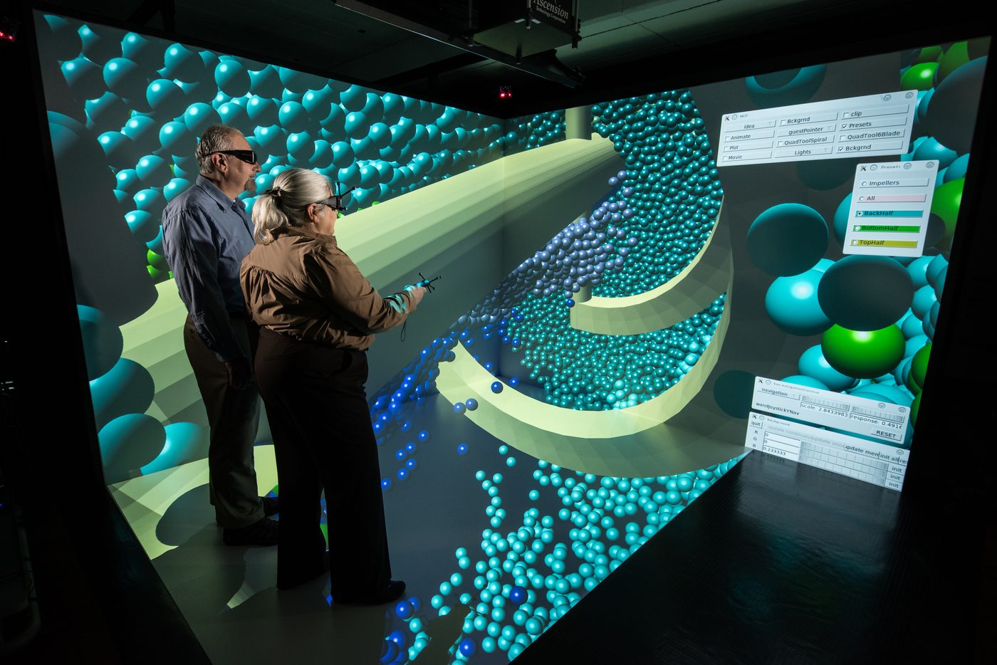 Two people wearing stereo glasses interact with a 3D particle simulation projected on the walls and floor of a CAVE immersive visualization room; one holds a tracked wand